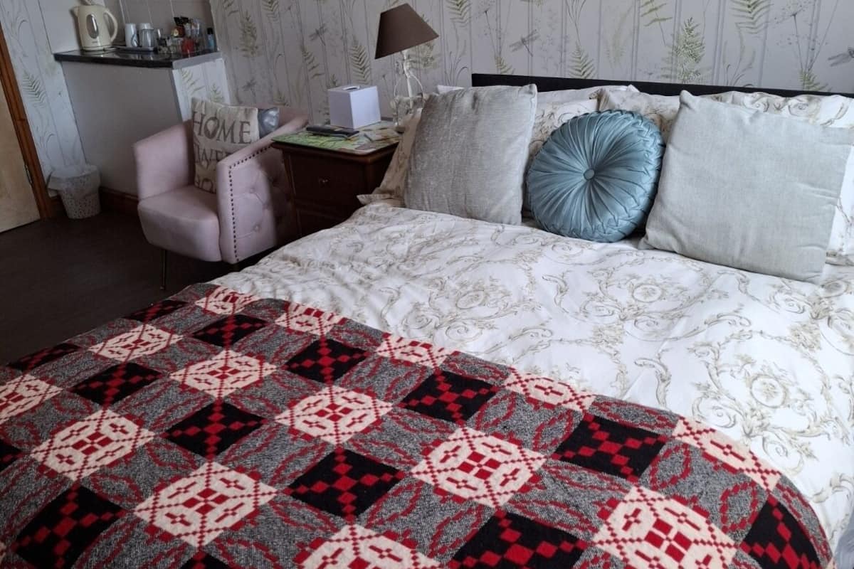 A neatly arranged bedroom with a patterned bedspread, a chair, a bedside table with a lamp, and a framed picture on the wall.