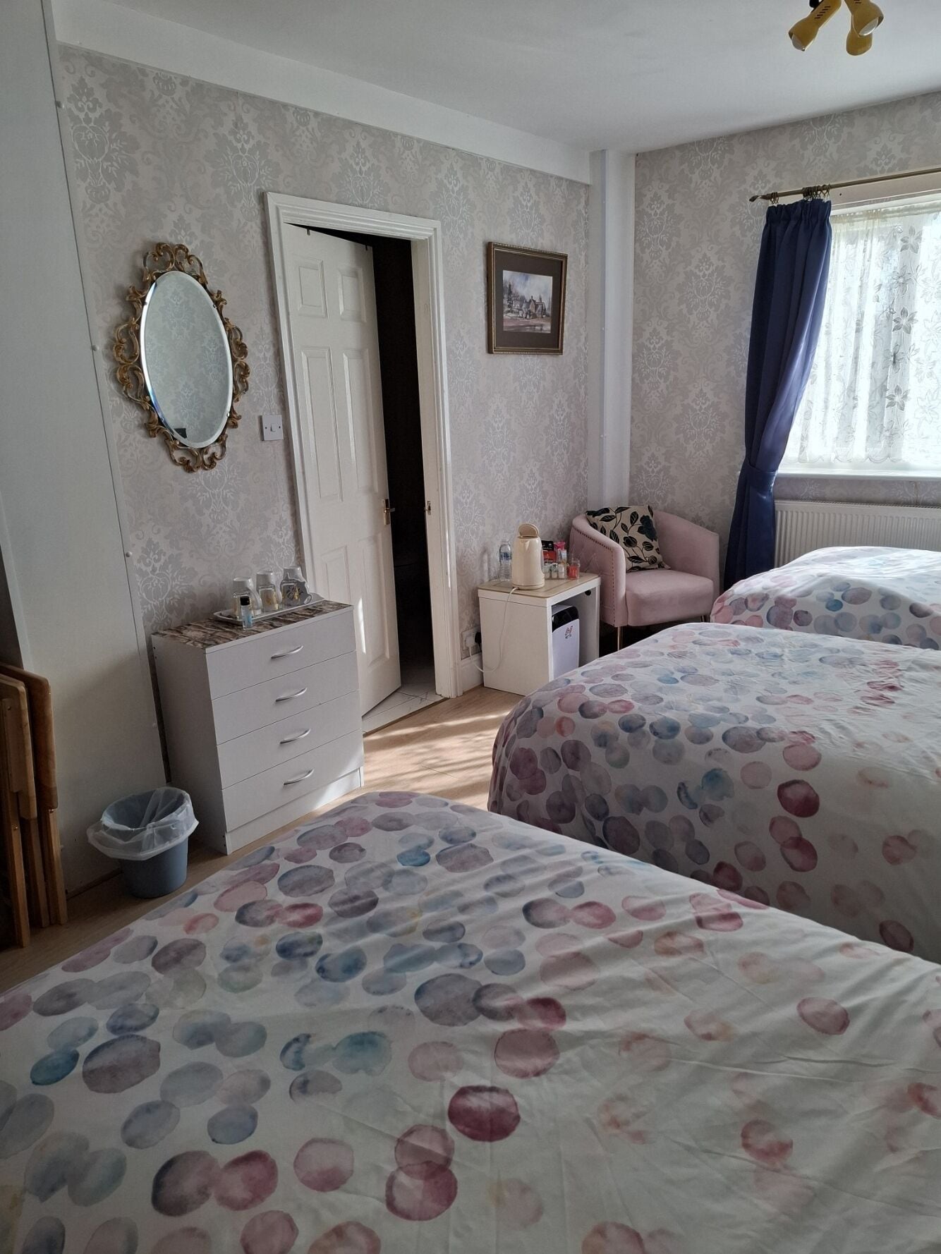 A bedroom with two beds, a dresser, a chair, a small table with items, and a door leading to another room.