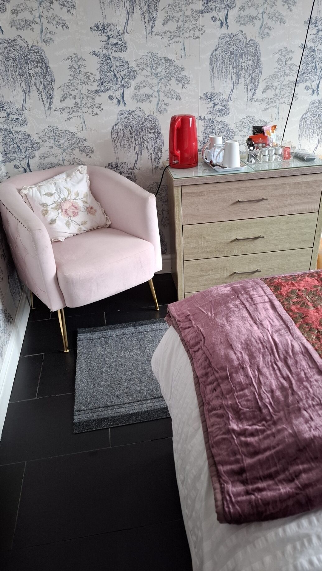 Makuuhuoneessa on vaaleanpunainen nojatuoli, puinen lipasto, punainen vedenkeitin ja sänky, jossa on vaaleanpunainen peitto.