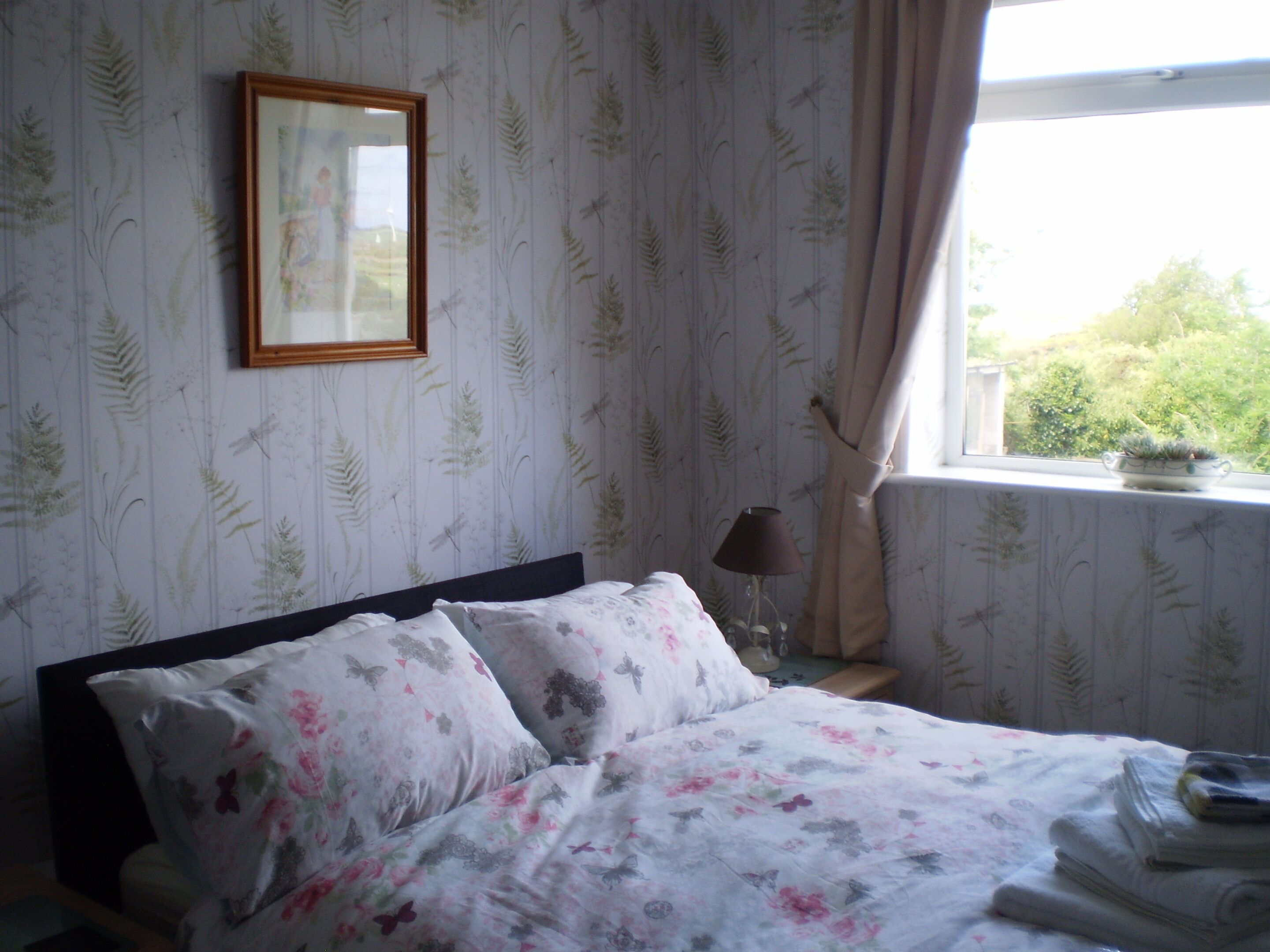 Une chambre à coucher avec un lit, une literie à motifs floraux, un tableau encadré au mur, une fenêtre avec des rideaux et une lampe de chevet.