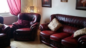 A living room with a dark red leather sofa and armchair, a red throw blanket, and a red curtain. There are three framed floral paintings on the wall.