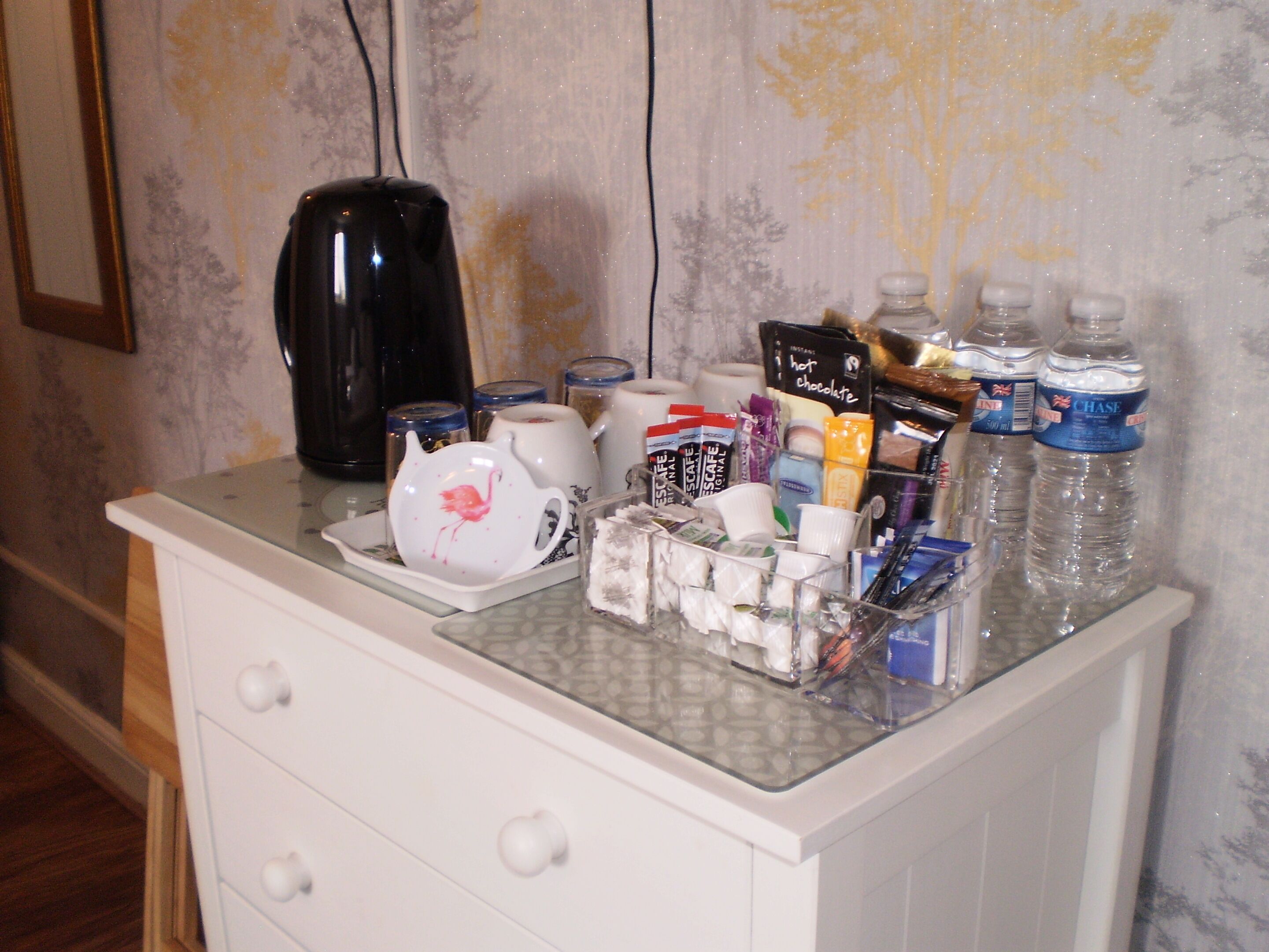 Un minibar con diversos artículos, como una cafetera, botellas de agua, aperitivos y un juego de té.