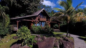 A wooden house with a balcony, surrounded by tropical vegetation and rocks. - Le Jardin Malanga (Trois-Rivières)