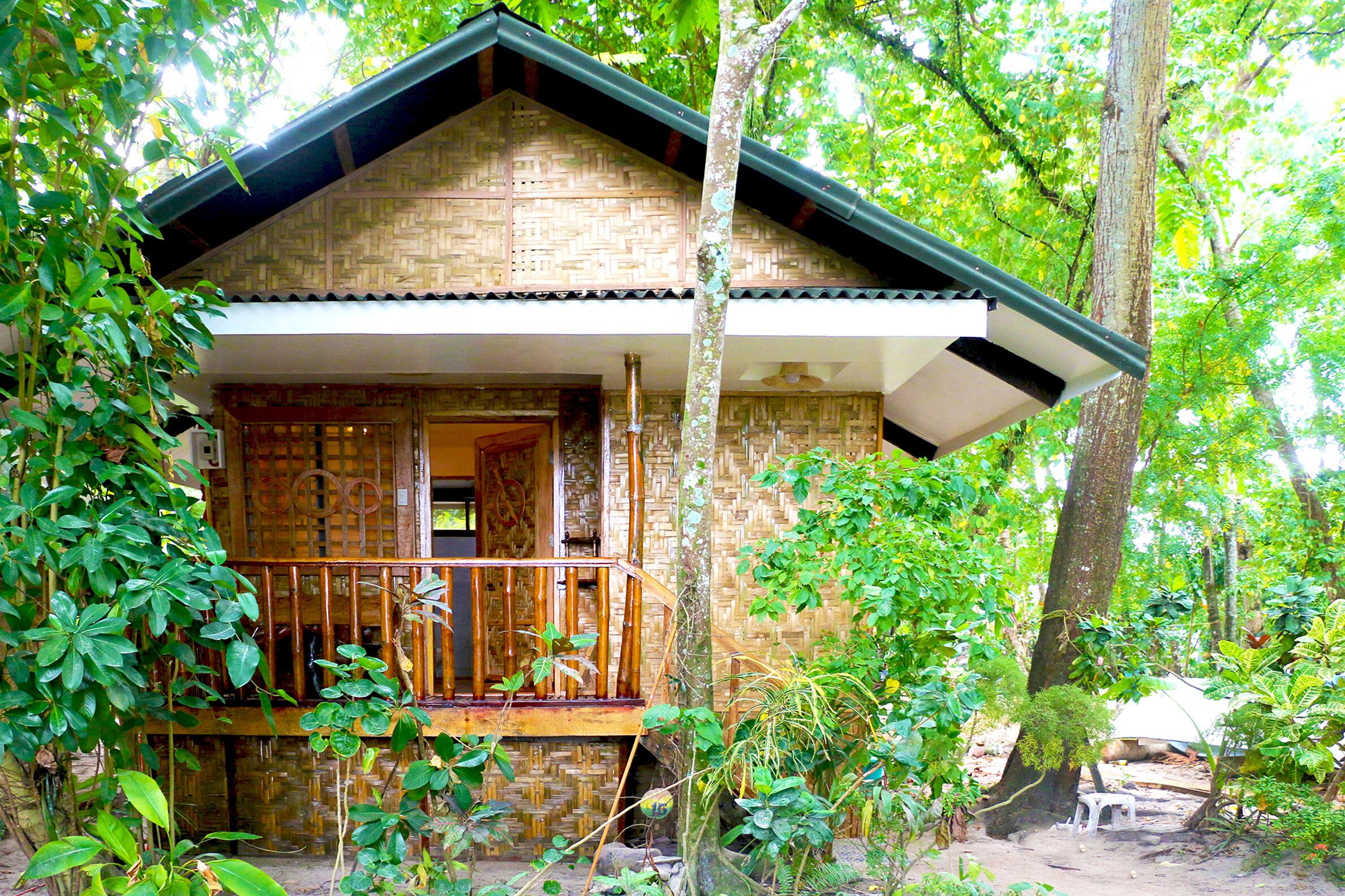 A rustic wooden house with a balcony, surrounded by lush greenery.