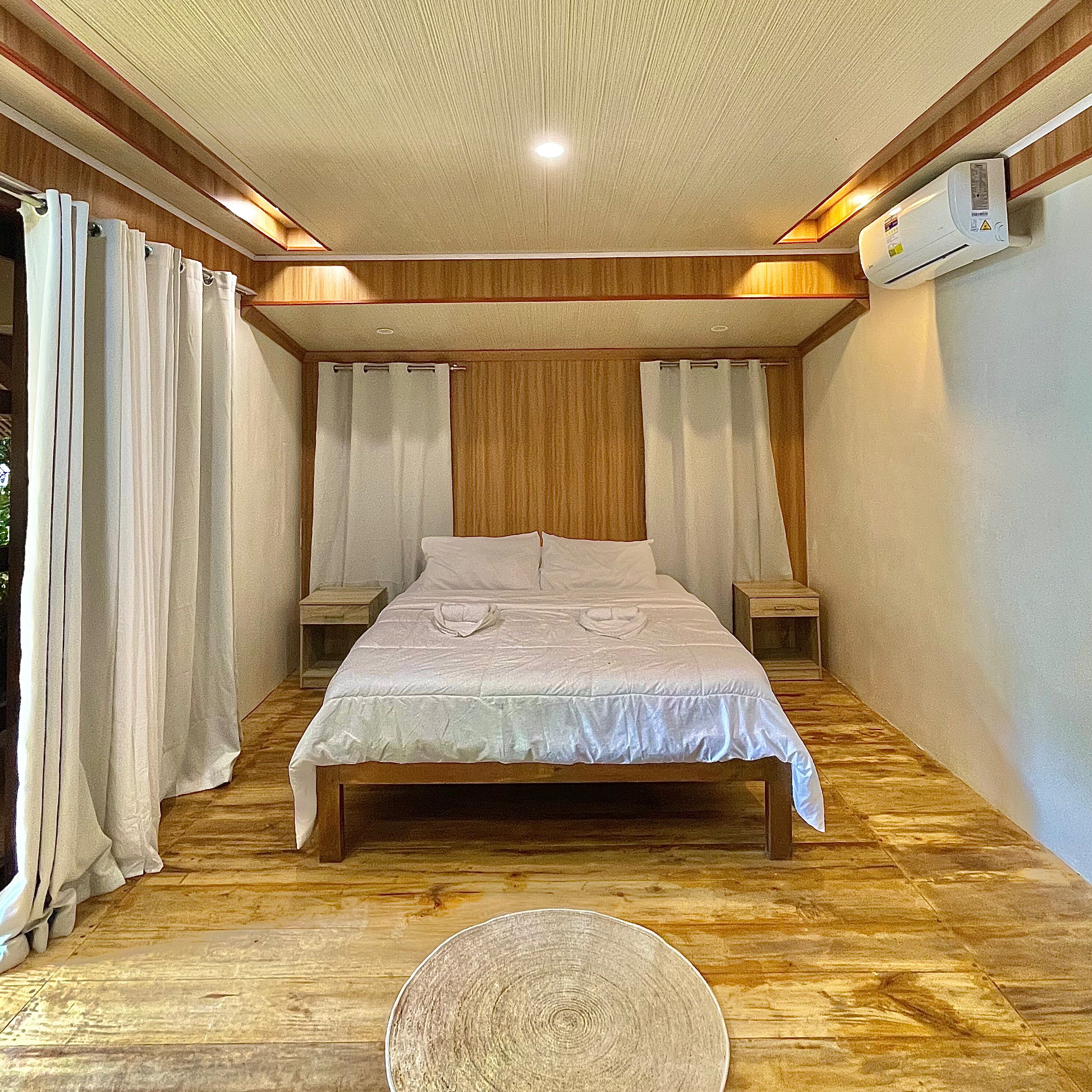 A bedroom with a wooden floor, a bed with white bedding, two bedside tables, and an air conditioning unit on the wall.