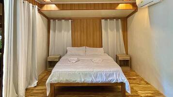 A bedroom with a wooden floor, a bed with white bedding, two bedside tables, and an air conditioning unit on the wall.