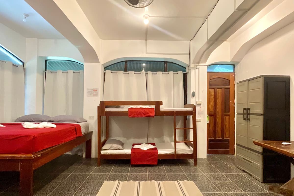 A hotel room with a bunk bed, a single bed, a fan, a small table, and a door leading to a kitchen area.