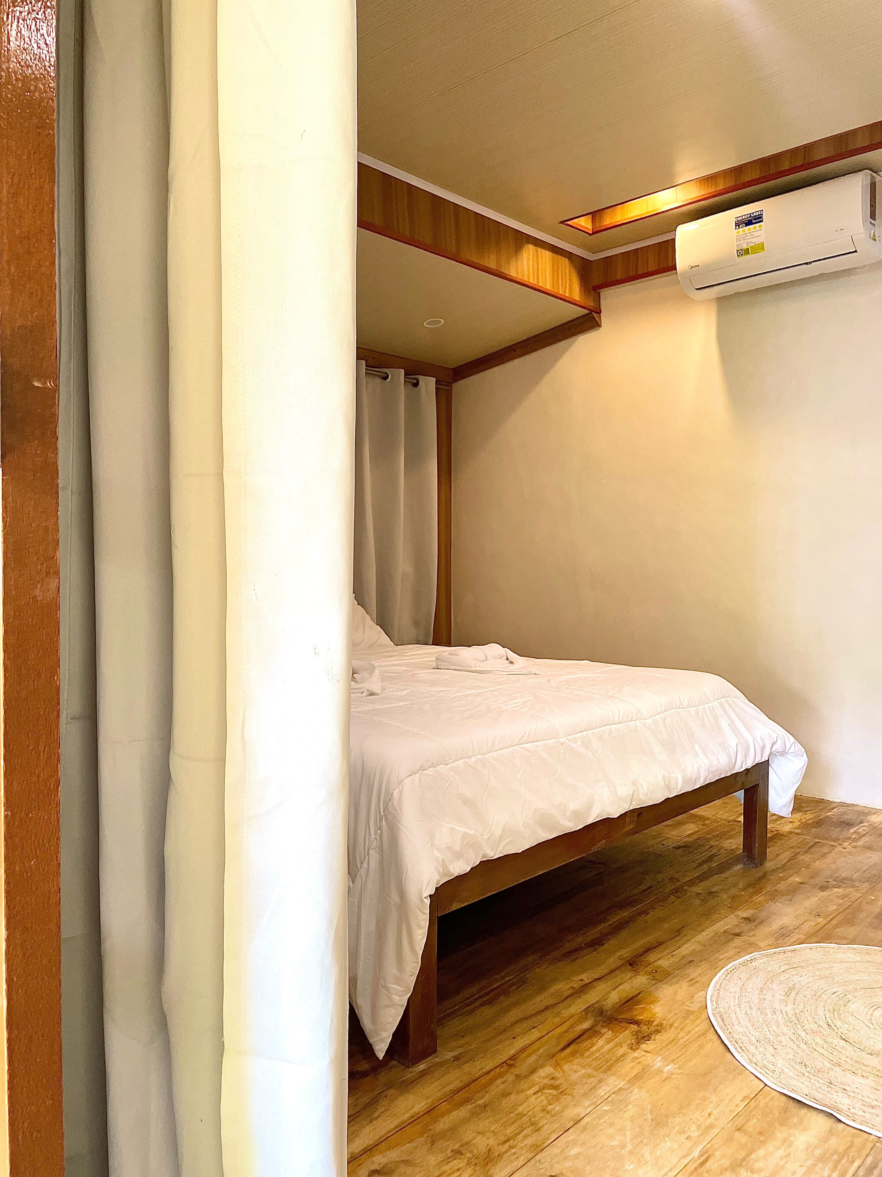 Una cama individual en una habitación con suelo de madera, cortina blanca y unidad de aire acondicionado en la pared.