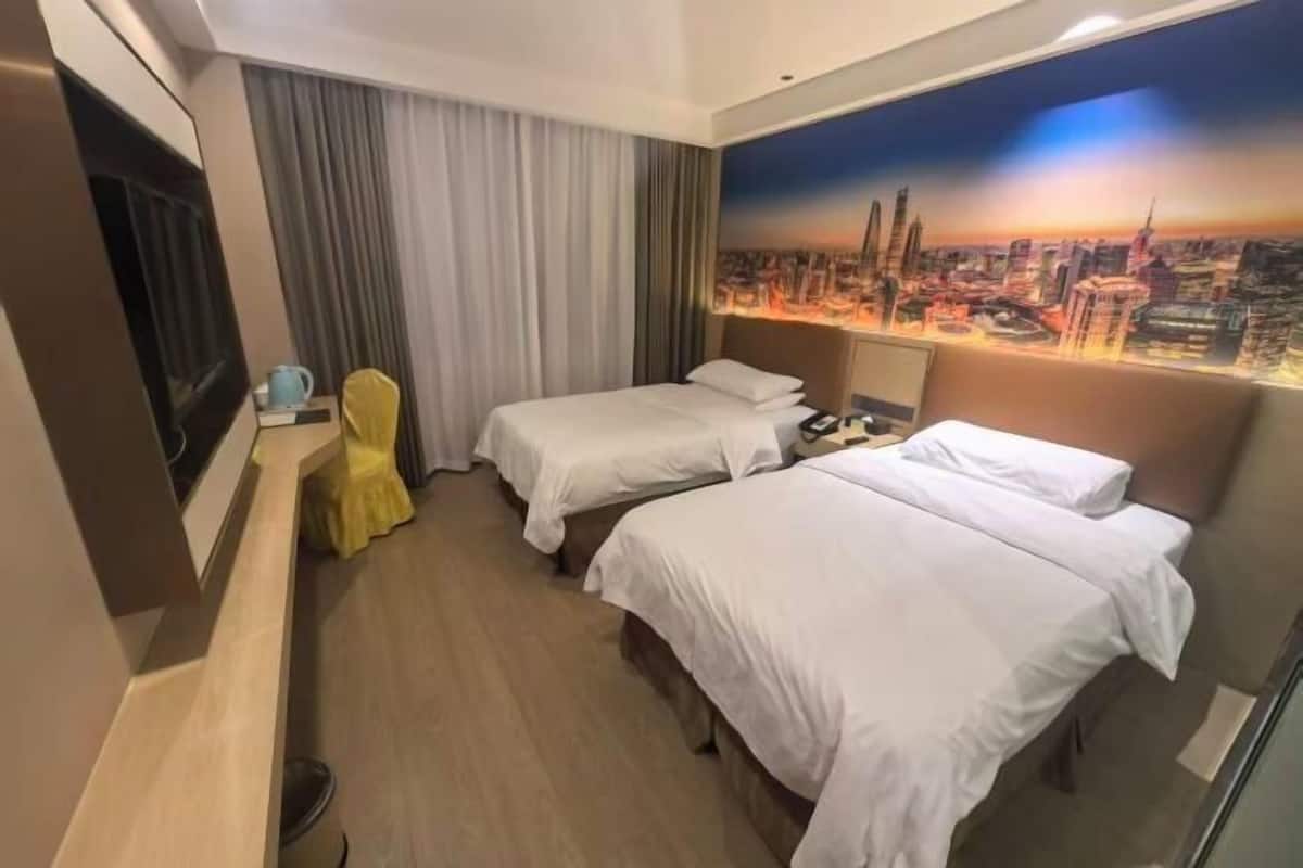 A hotel room with two beds, a TV, a desk with a chair, and a cityscape mural on the wall.
