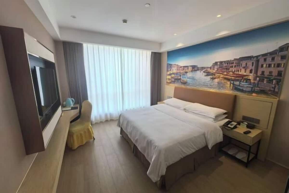 A hotel room with a bed, a desk with a chair, a TV, a nightstand, and a painting of a cityscape.
