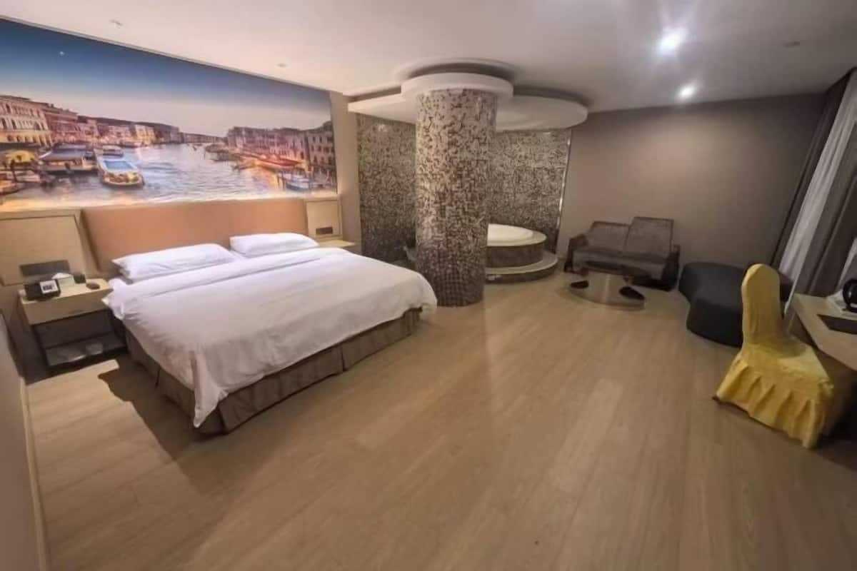 A hotel room with a bed, a desk, a chair, a large window with a cityscape view, and a decorative column.