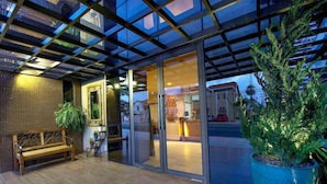 A modern building entrance with a glass door, a wooden bench, and a large potted plant. - Carmel Express Hotel (FORTALEZA)