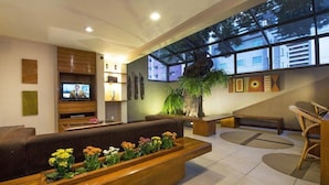 A modern living room with a sofa, a wooden coffee table with flowers, a television, and a large window with a view of buildings. - Carmel Express Hotel (FORTALEZA)