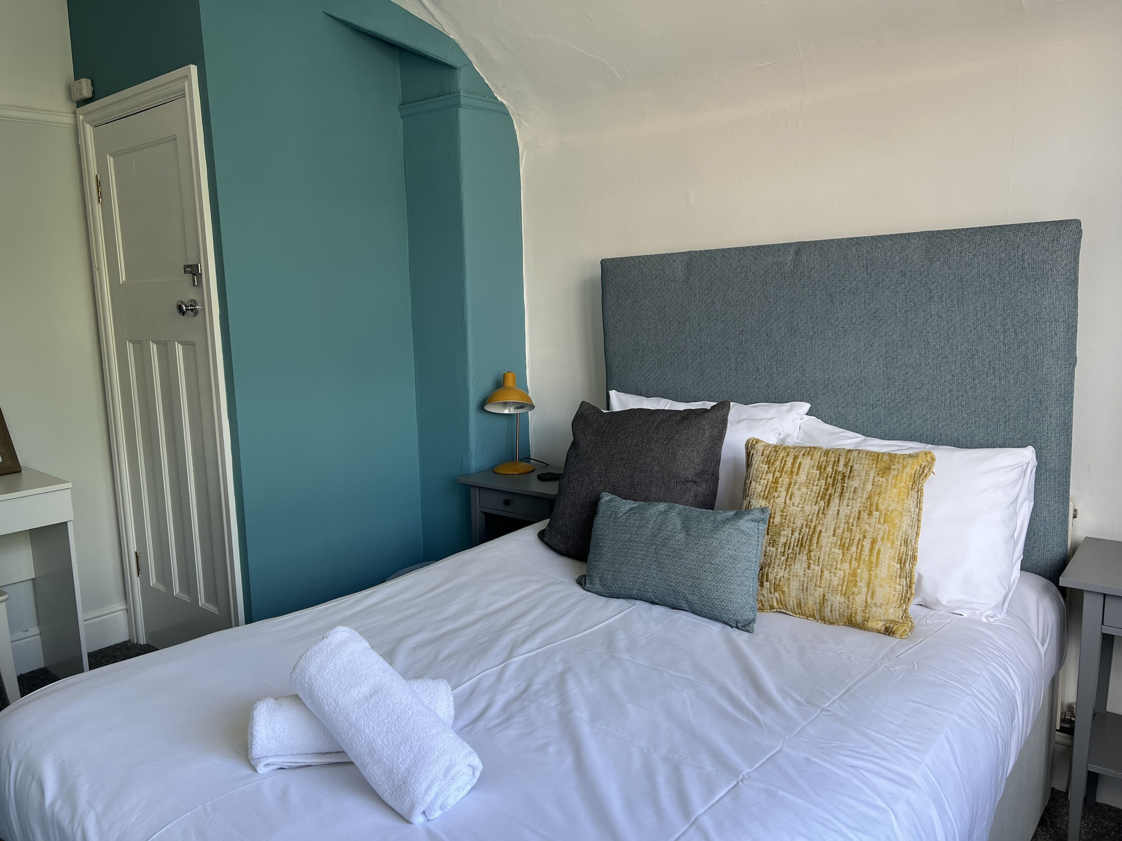 A neatly made bed with white linens and a rolled white towel, a grey headboard, and a bedside table with a lamp and a small drawer.