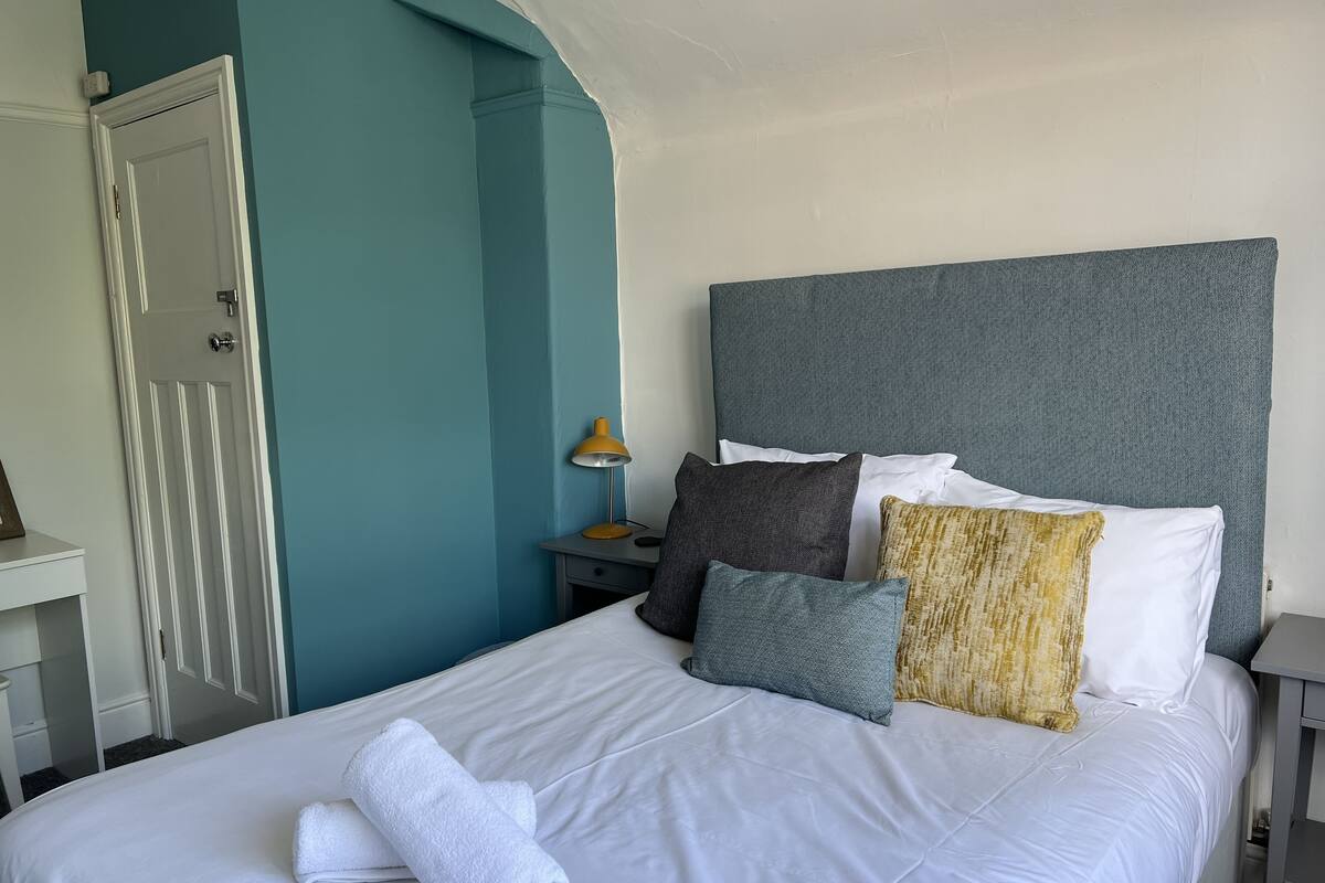 A neatly made bed with white linens and a rolled white towel, a grey headboard, and a bedside table with a lamp and a small drawer.