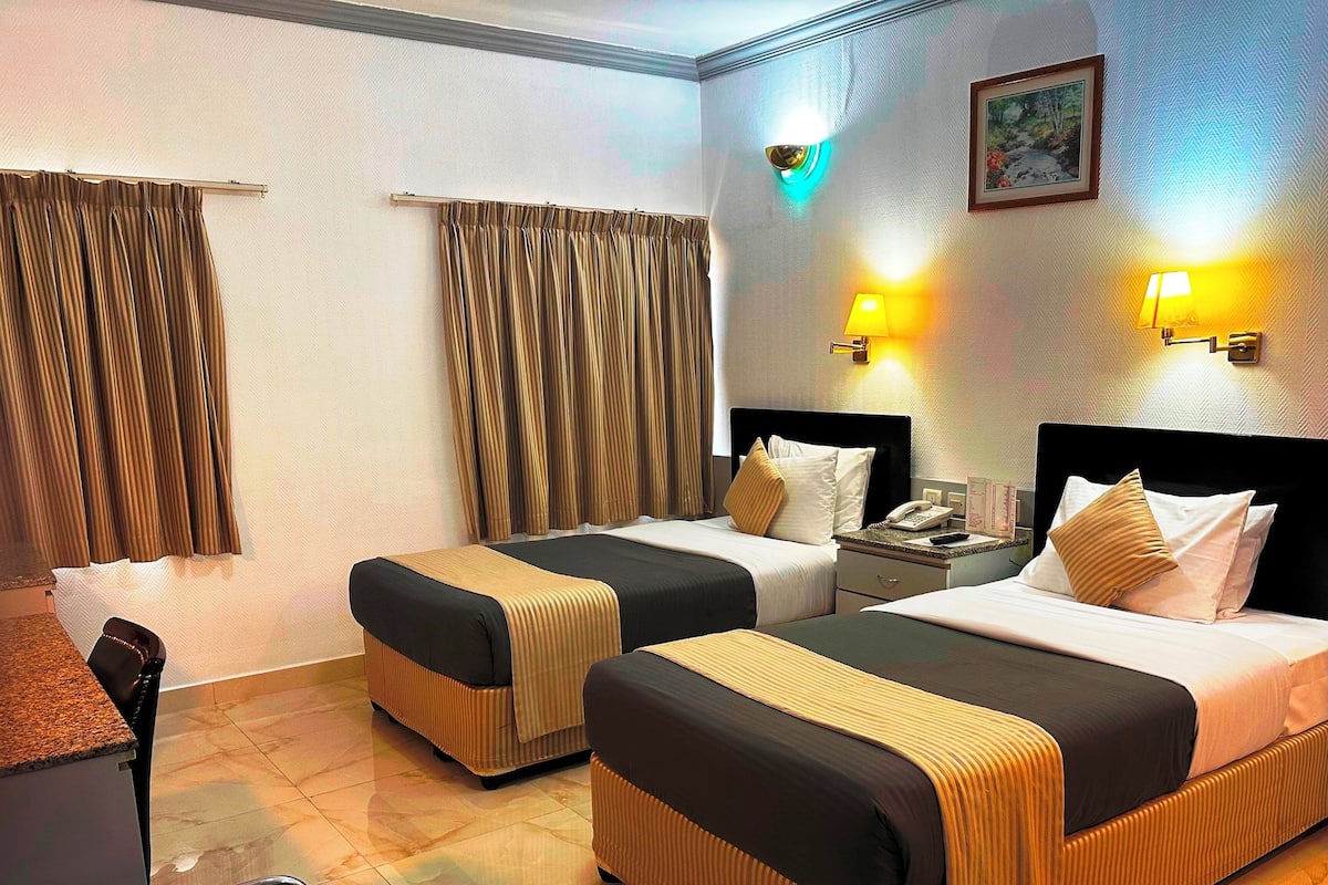 Une chambre d’hôtel avec deux lits, un bureau et une chaise. On y trouve des rideaux, des appliques murales et un tableau encadré accroché au mur.
