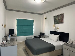 Een hotelkamer met een bed, een bureau, een stoel, een raam met groene luiken, een schilderij aan de muur en een telefoon op het bureau.