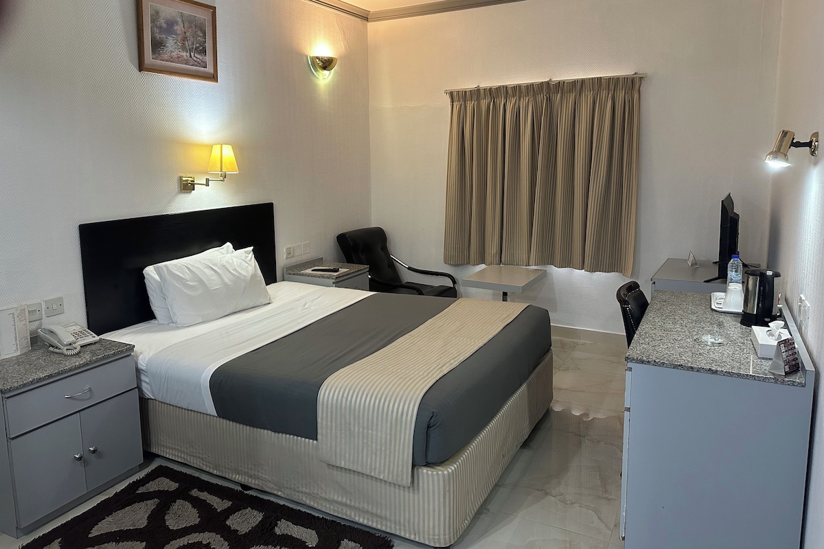 Habitación de hotel con cama, mesita de noche, silla, escritorio y una pequeña área de kitchenette.