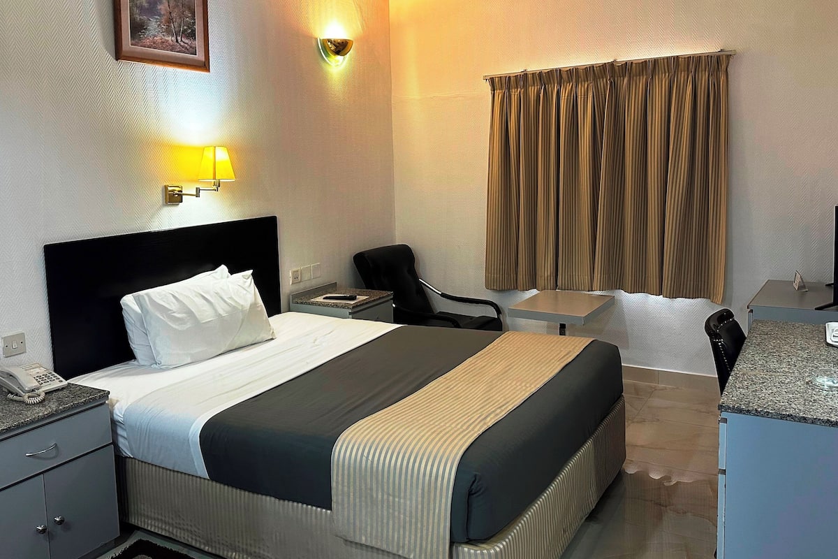 A hotel room with a bed, bedside table, chair, desk, and a window with curtains.