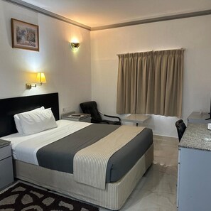 A hotel room with a bed, a desk, a chair, a small table, and a window with curtains.
