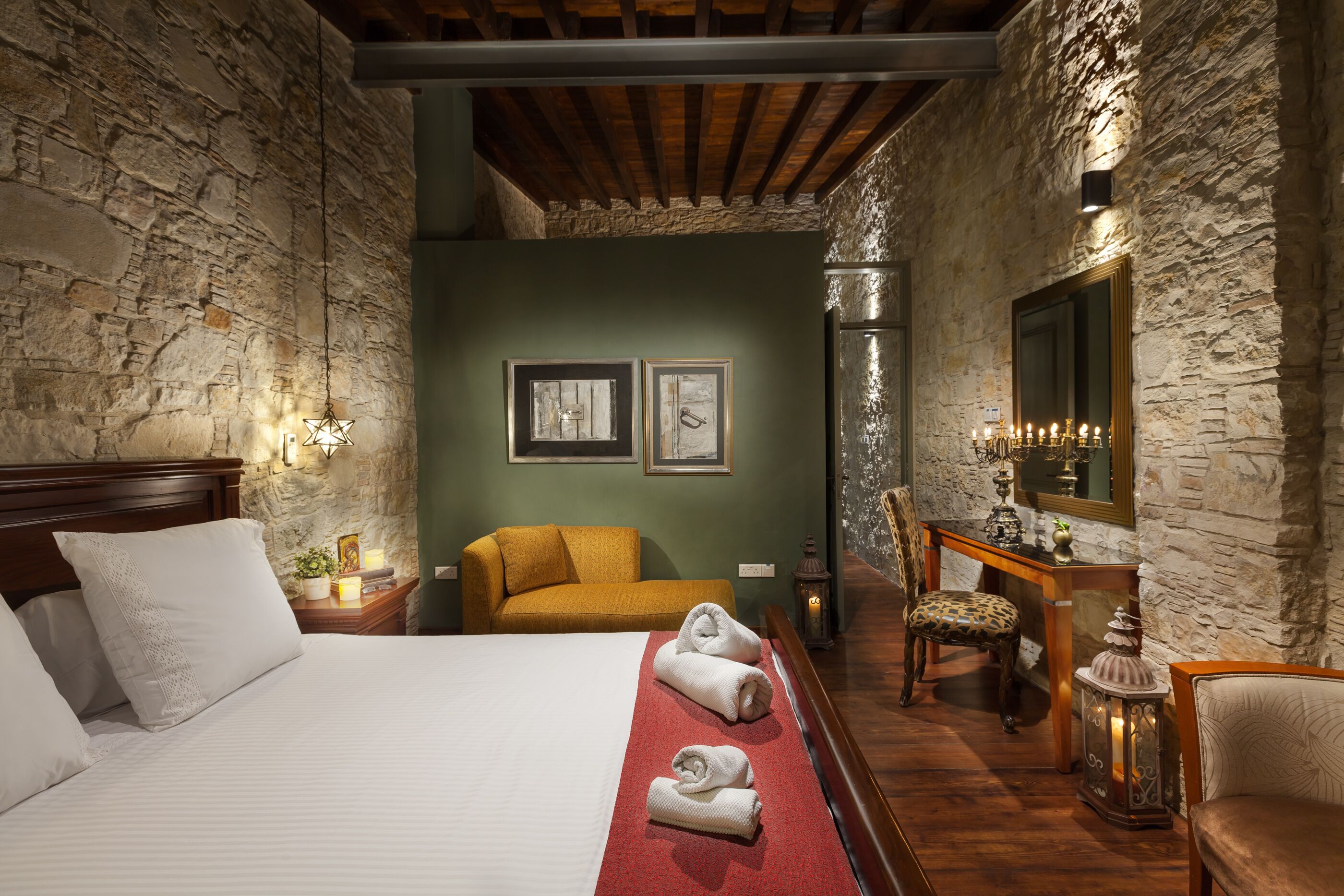 A hotel room with a stone wall, a wooden bed, a sofa, a chair, and a desk with a mirror.