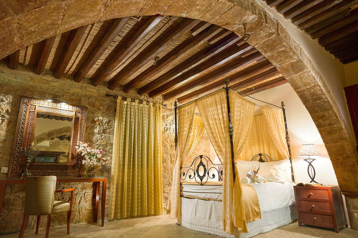 A bedroom with a four-poster bed, a wooden chair, a nightstand, and a mirror on the wall.