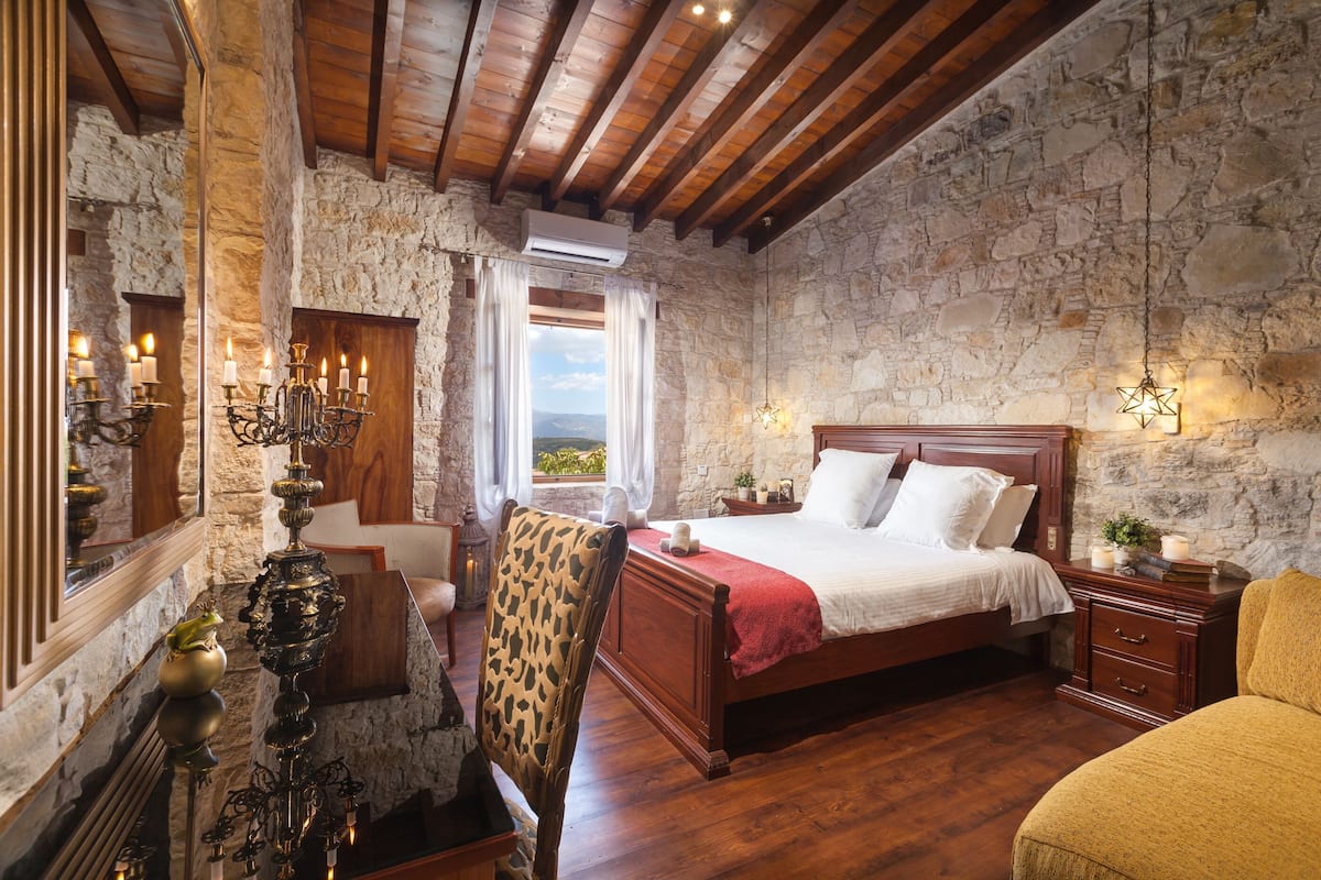 A bedroom with a stone wall, wooden ceiling beams, a bed with white linens, a nightstand, and a chair.