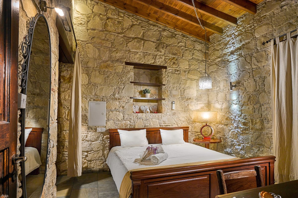 A stone-walled bedroom with a wooden bed, a bedside table with a lamp, and a small shelf with decorative items.