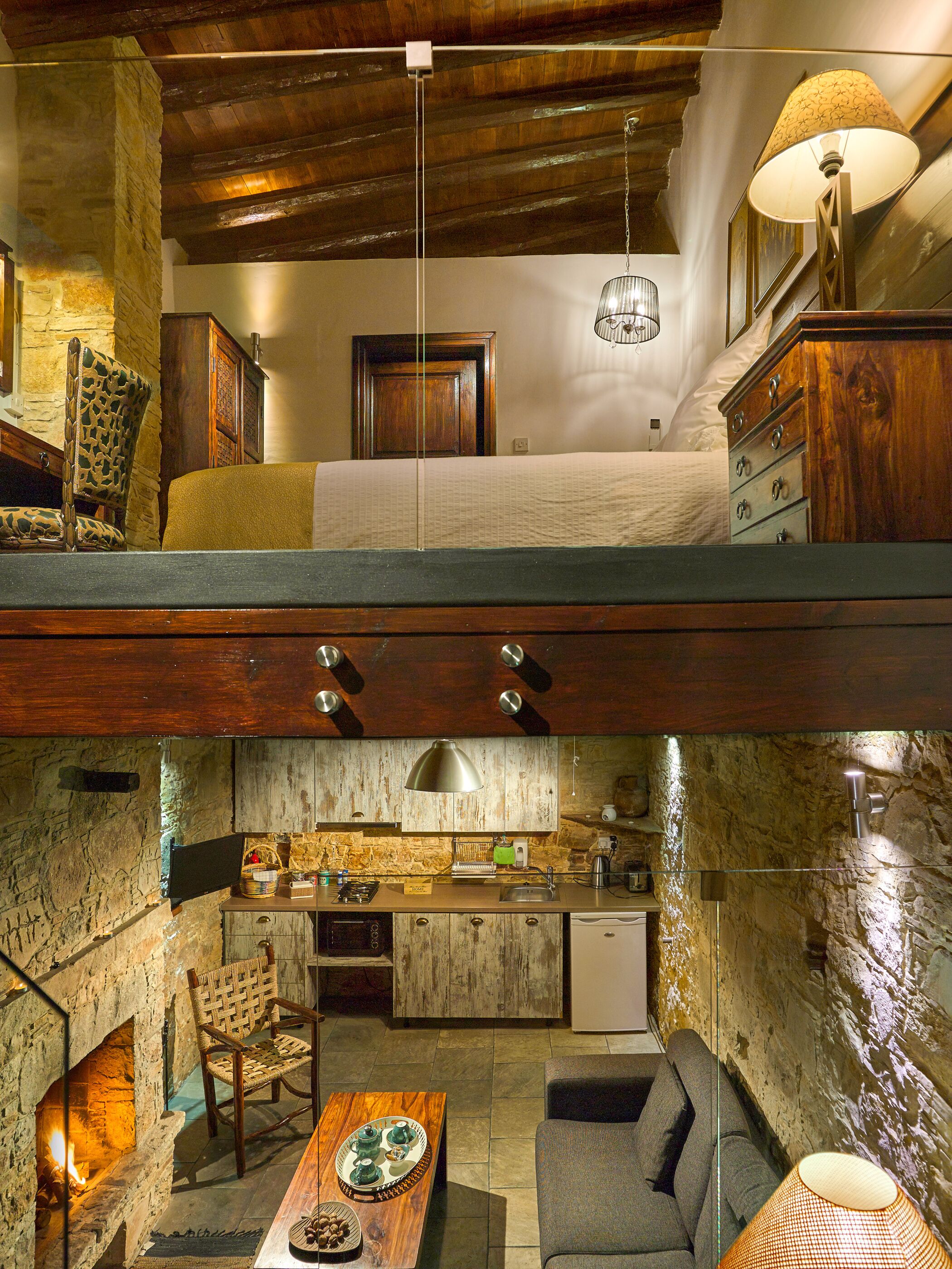 A rustic interior with a loft bedroom, a stone fireplace, a kitchen with a stone wall, and a dining area with a wooden table.