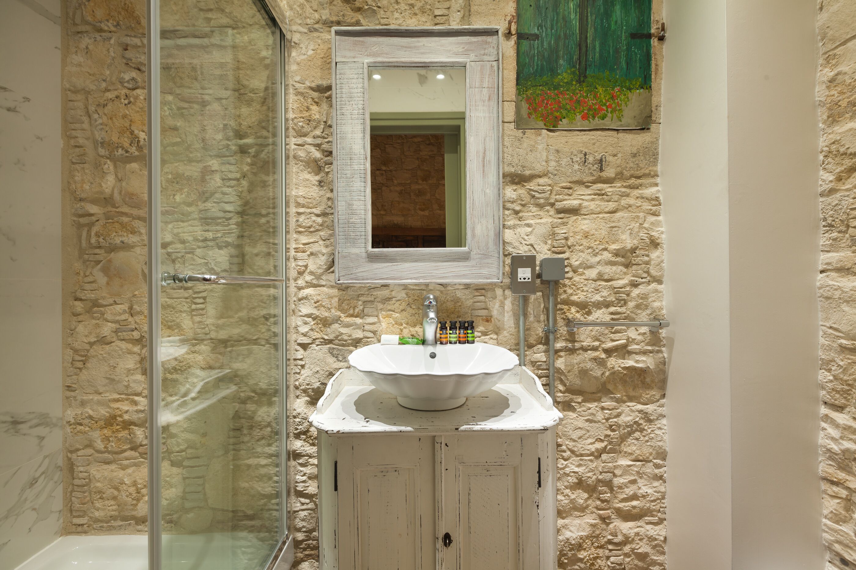 A bathroom with a stone wall, a glass shower enclosure, a white sink with a mirror above it, and a green door.