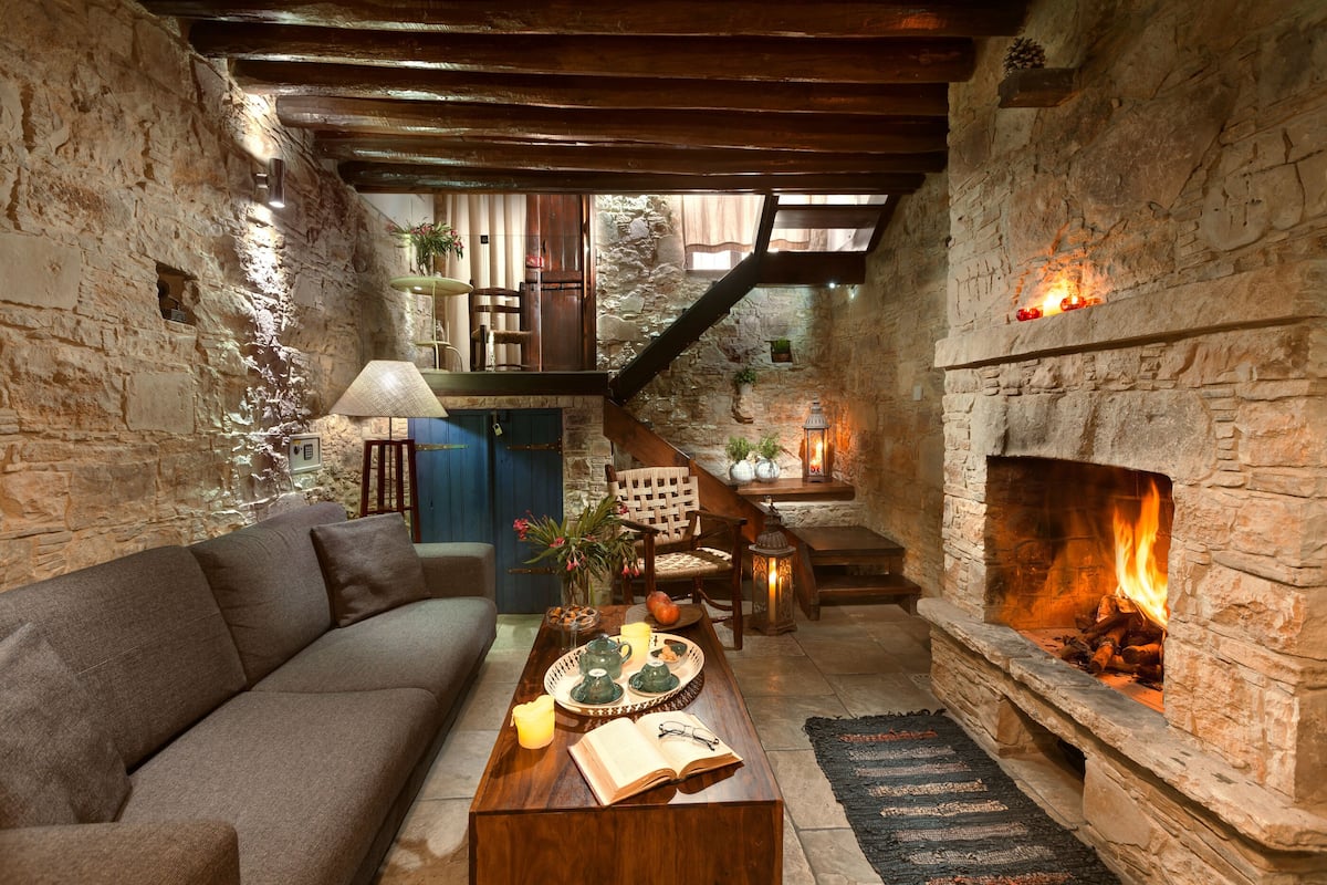 A cozy room with a stone fireplace, a sofa, a wooden coffee table with a book and teapots, and a staircase leading to an upper level.