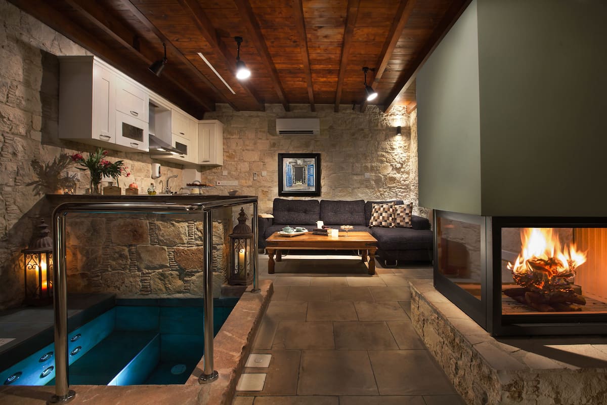 A stone-walled room with a wooden ceiling, a built-in fireplace, a sofa, a coffee table, and a stone tub.