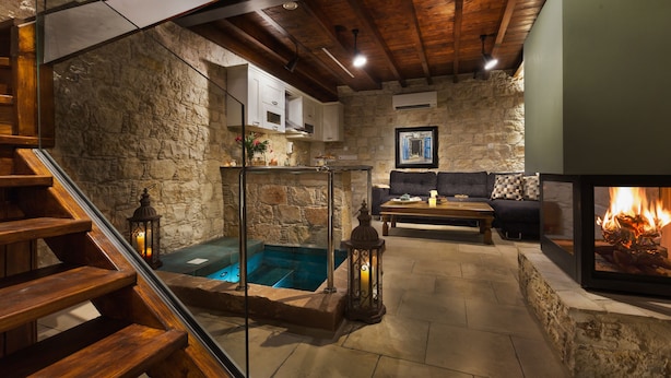 A stone-walled room with a wooden staircase, a glass-enclosed pool, a fireplace, and a sofa with cushions.
