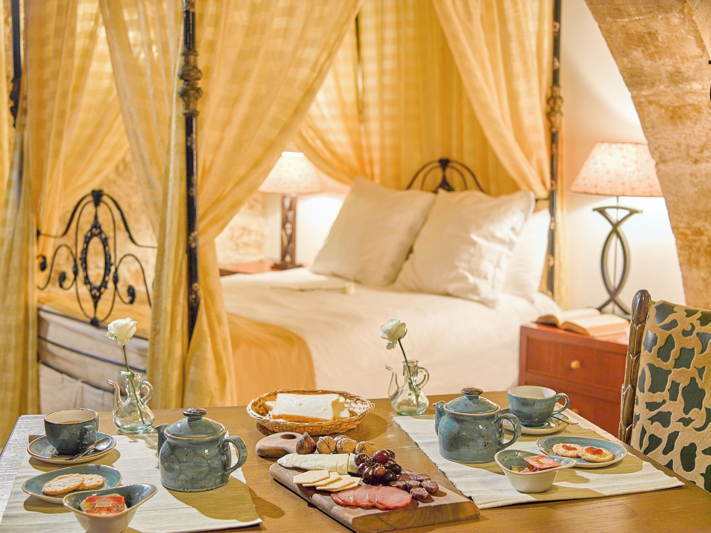 Une chambre à coucher avec un lit à baldaquin, des tables de chevet avec des lampes, une table pour le petit-déjeuner avec un service à thé, et une planche de fromages et de fruits.