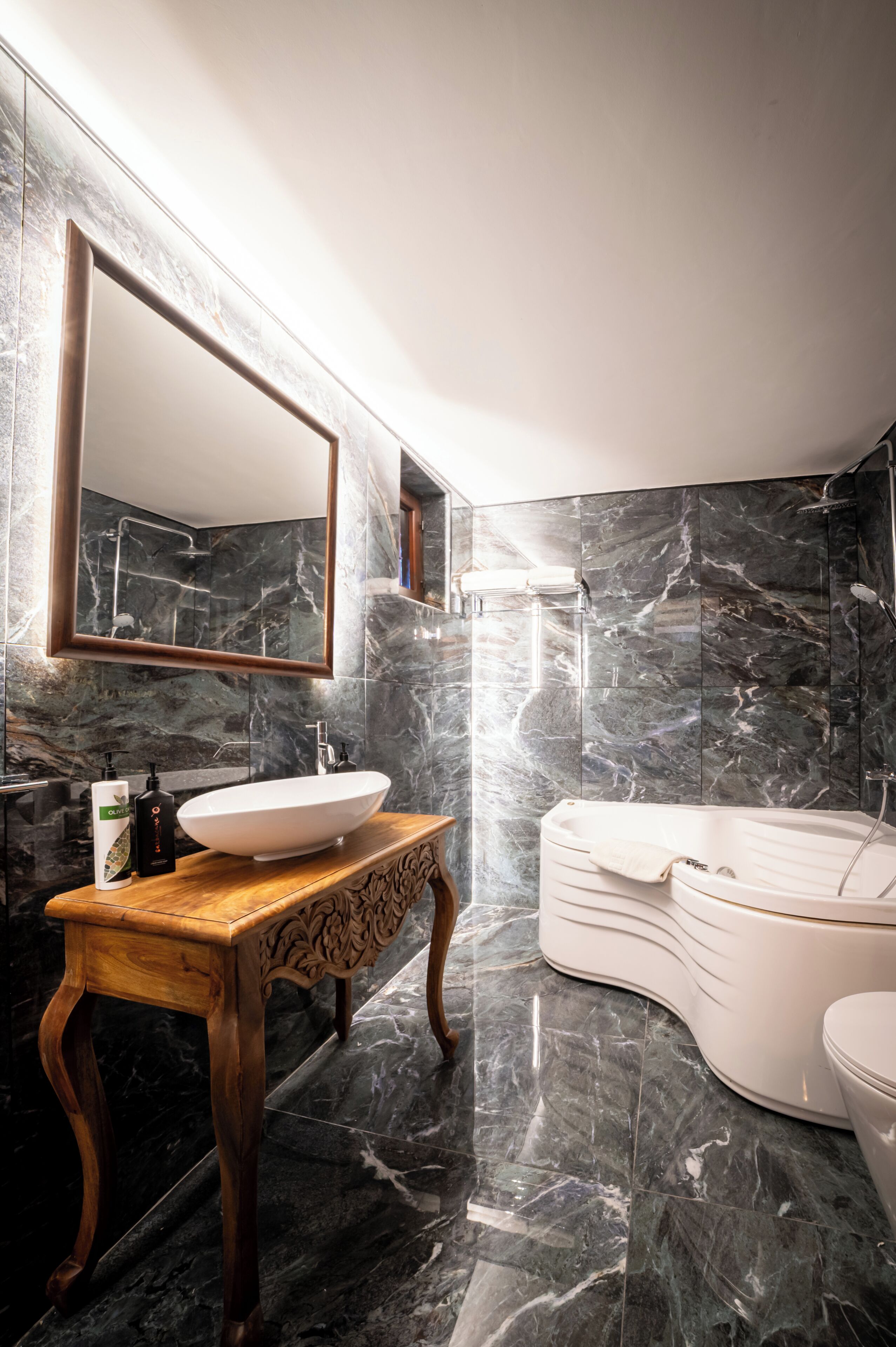A modern bathroom with a large mirror, a wooden vanity, a white sink, a bathtub, and marble walls.