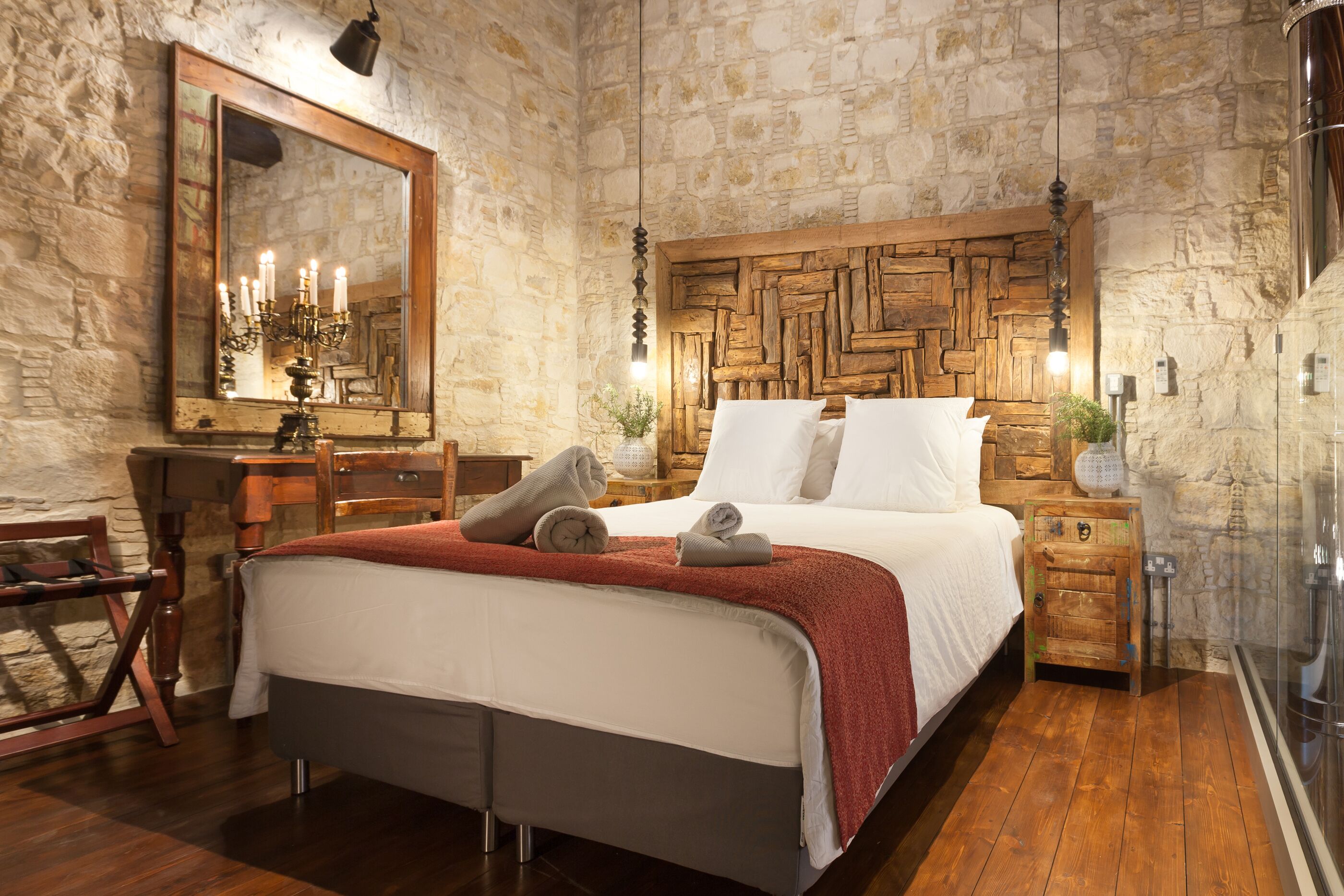 Un dormitorio con pared de piedra, cabecero de madera, cama con ropa blanca, mesita de noche de madera y un espejo.