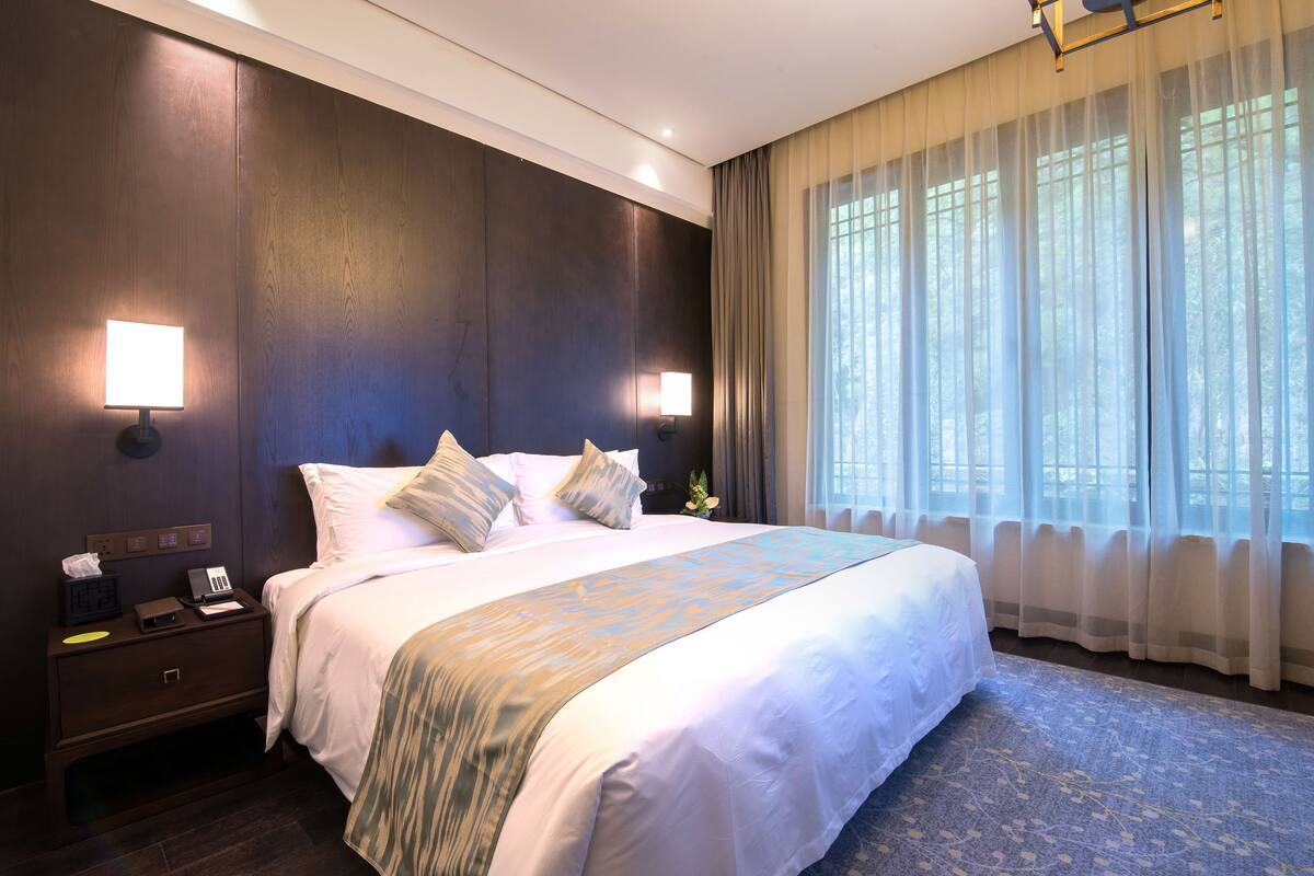 Een hotelkamer met een groot bed, twee bedlampjes, een nachtkastje en een raam met dunne gordijnen.