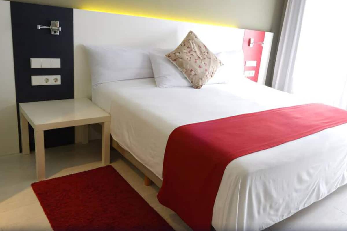 A hotel room with a bed, a nightstand, a red rug, and a window with curtains.