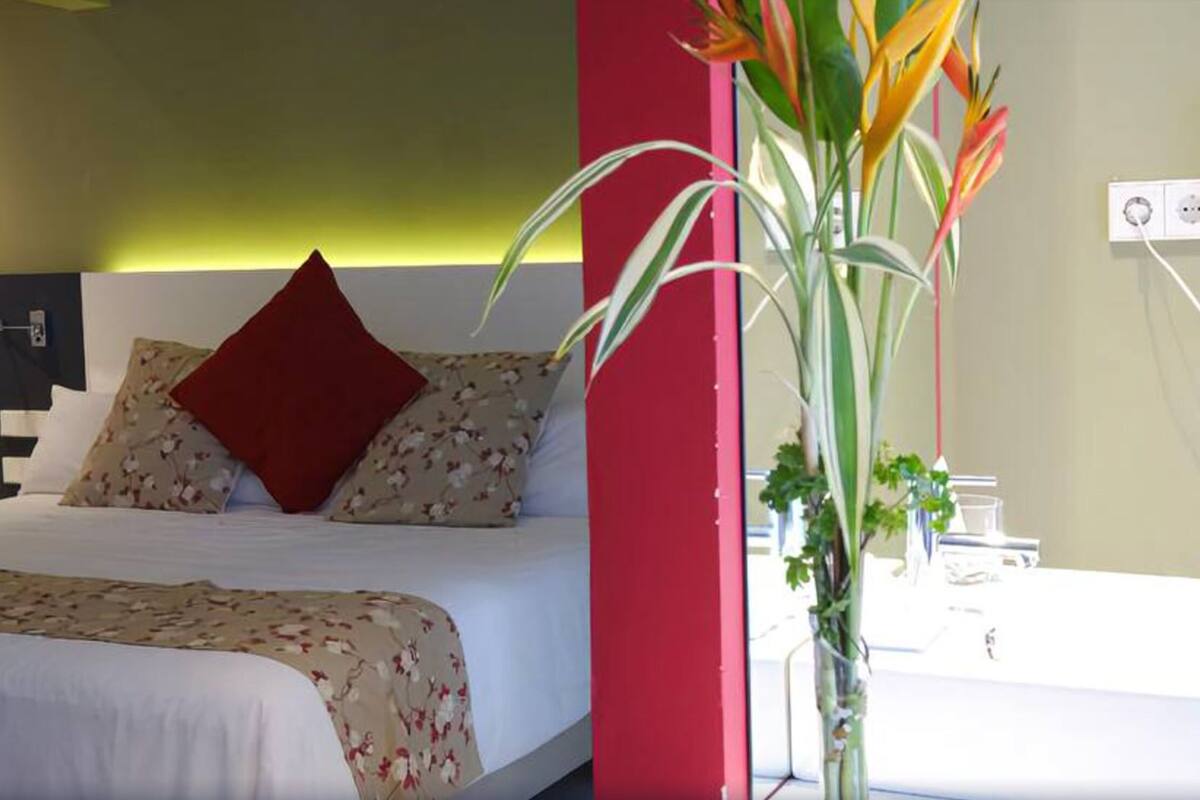 A hotel room with a bed, a red accent wall, a bedside table with a lamp, and a plant.