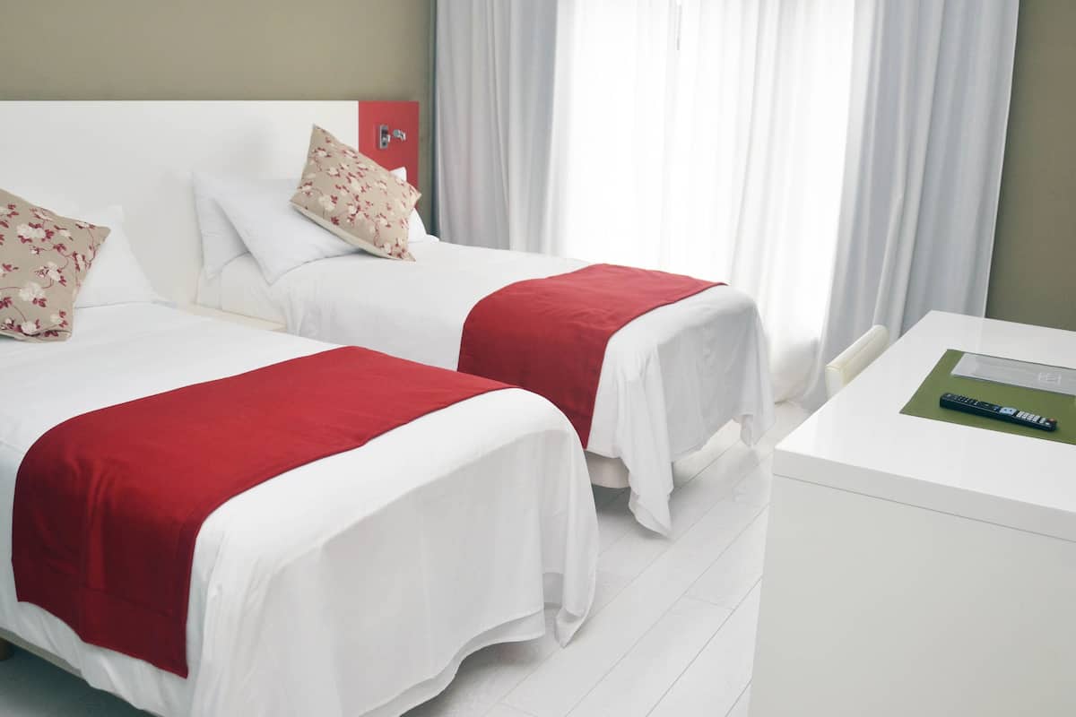 A hotel room with two beds, each with white bedding and red accents, a small table with a remote control, and a red door.