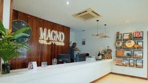 A reception desk with a wooden panel and a clock, a person behind the desk, and a wall with framed pictures and awards. - Magno Suites (Malabo)