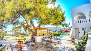 An outdoor seating area with stone flooring, tables, and chairs, a bar, and a large tree providing shade. - Casa Grande (Mykonos)