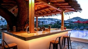An outdoor bar area with a wooden structure, bar stools, and a view of a pool and buildings in the background. - Casa Grande (Mykonos)