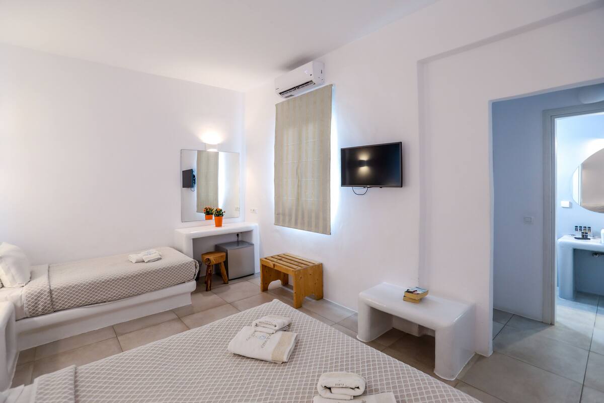 A modern hotel room with two single beds, a small wooden stool, a bench, a TV mounted on the wall, and a bathroom visible through an open door.