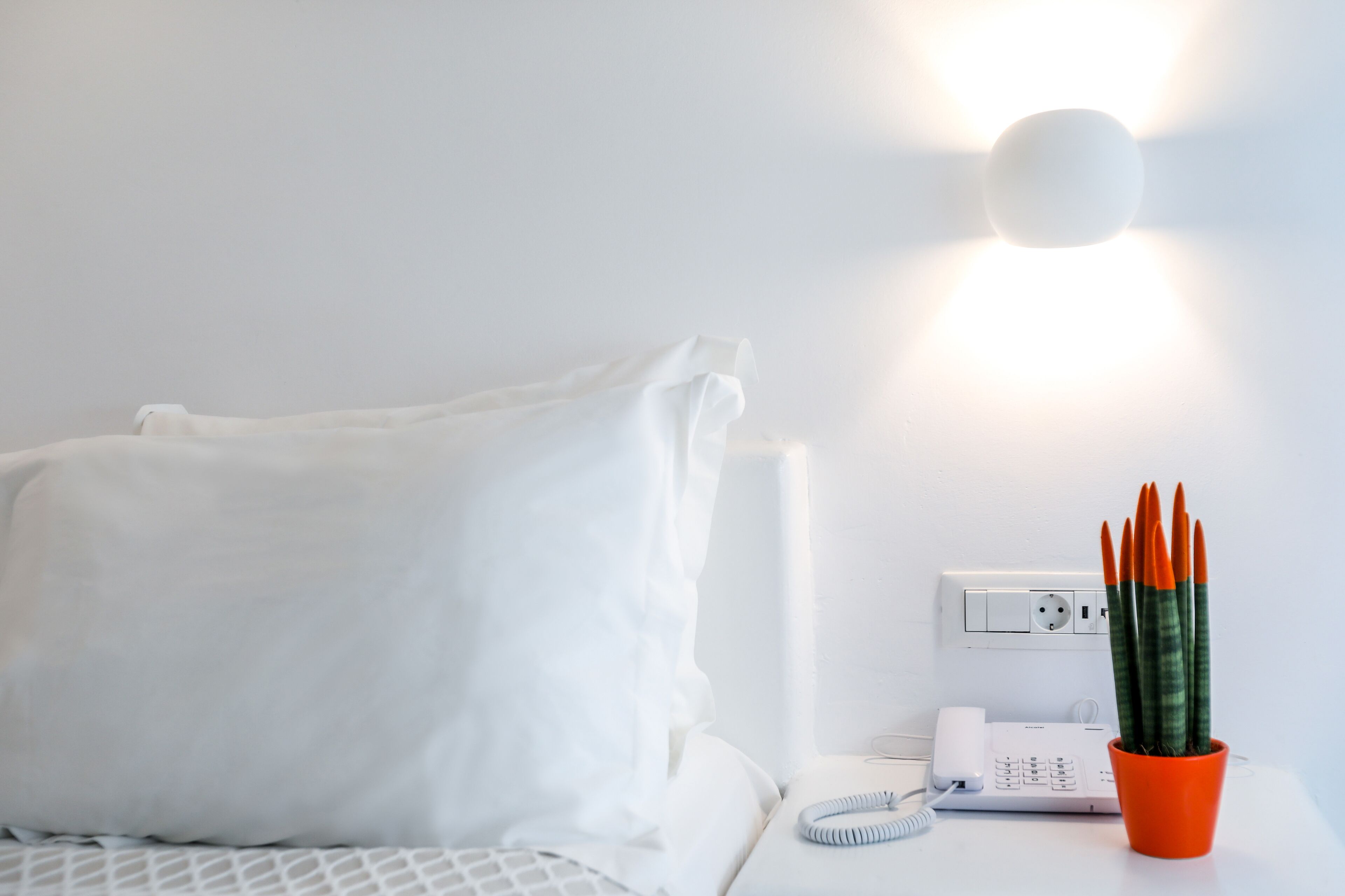 A white bed with a white pillow, a white bedside table with a telephone, a red pot with a plant, and a wall socket.
