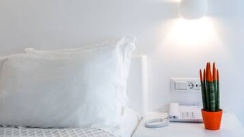 A white bed with a white pillow, a white bedside table with a telephone, a red pot with a plant, and a wall socket.