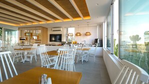 A modern dining area with white chairs and wooden tables, a kitchen in the background, and large windows with a view. - Casa Grande (Mykonos)