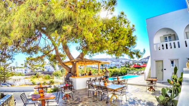 An outdoor patio with stone flooring, wooden tables and chairs, a bar area, and a view of a white building and a pool. - Casa Grande (Mykonos)