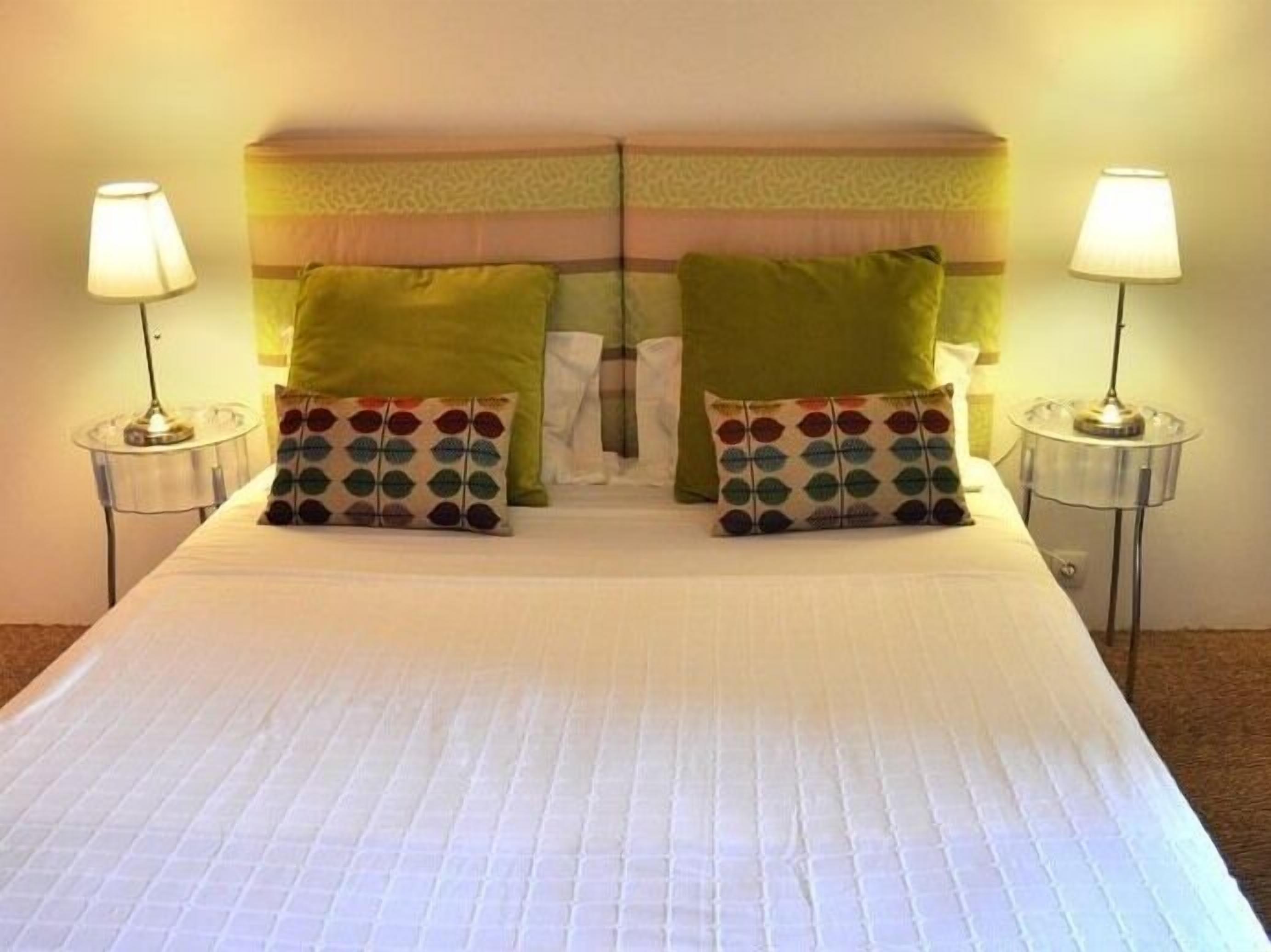 Un lit bien fait, recouvert d’une couette blanche, avec deux oreillers verts et un oreiller à motifs posé sur la tête de lit. Deux lampes de chevet sont placées de chaque côté du lit.