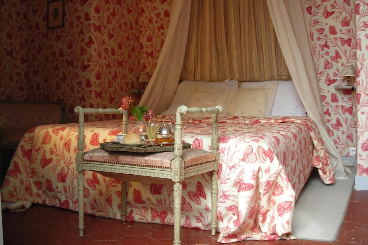 Une chambre à coucher avec un lit à baldaquin, un papier peint à motifs floraux et une table de chevet sur laquelle se trouve un plateau avec divers objets.